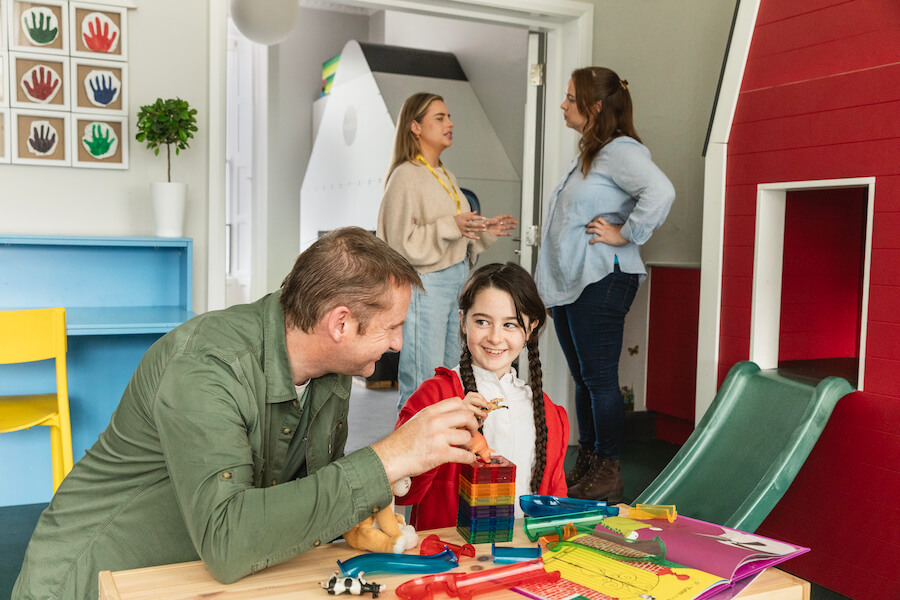 A father and daughter playing games while the mother talks with a woman more seriously